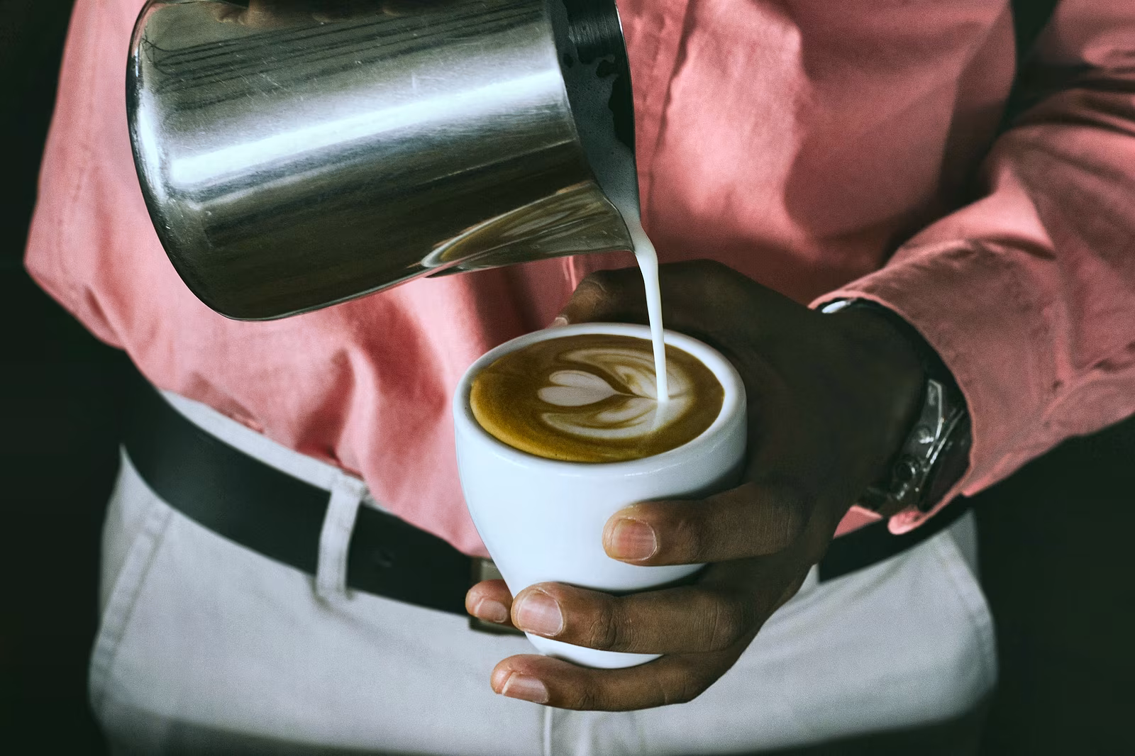 Milk from a metal pitcher being poured into a cup of coffee.